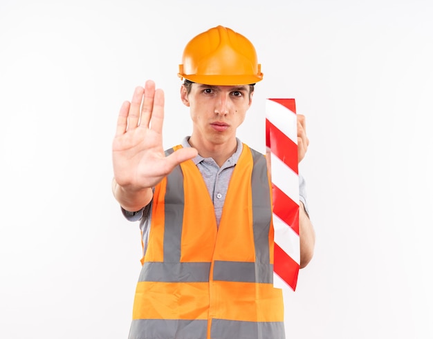 Strict young builder man in uniform holding duct tape showing stop gesture isolated on white wall