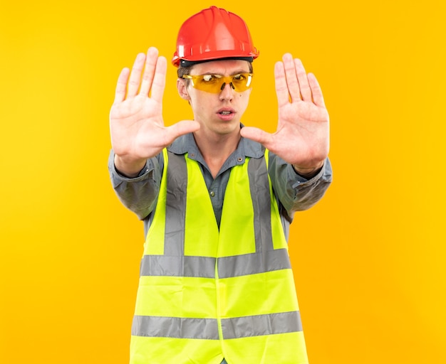 Strict young builder man in uniform wearing glasses showing stop gesture 