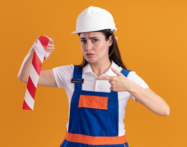 Rigorosa giovane donna costruttore in tenuta uniforme e punta al nastro adesivo isolato su parete arancione orange