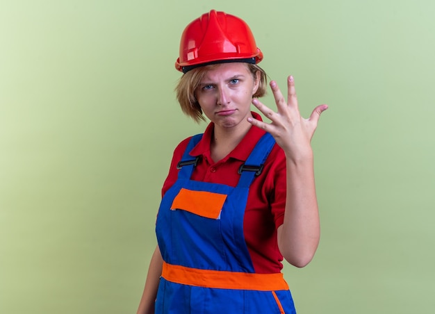 strict young builder woman in uniform showing five isolated on olive green wall