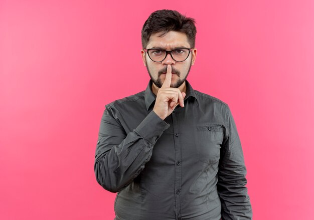 Strict young businessman wearing glasses showing silence gesture isolated on pink