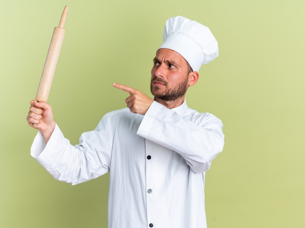 Strict young caucasian male cook in chef uniform and cap holding looking and pointing at rolling pin isolated on olive green wall