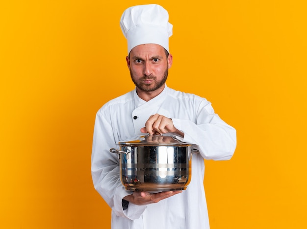 Rigoroso giovane maschio caucasico cuoco in uniforme da chef e cappuccio che tiene la pentola afferrando il coperchio