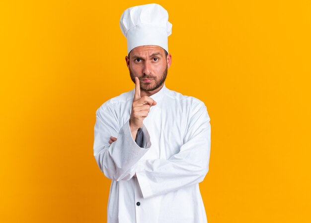 Строгий молодой кавказский повар в униформе шеф-повара и поднимающий кепку палец