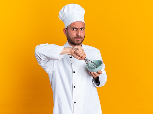 Строгий молодой кавказский повар в униформе шеф-повара и кепке взбивает яйцо в миске