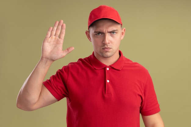 Strict young delivery man wearing uniform and cap showing stop gesture isolated on olive green wall