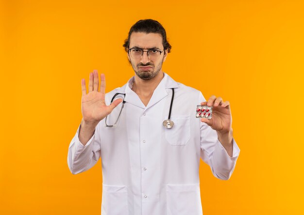Strict young doctor with medical glasses wearing medical robe with stethoscope holding pills and showing stop gesture on yellow