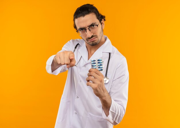 Strict young doctor with medical glasses wearing medical robe with stethoscope holding pills and showing you gesture on yellow