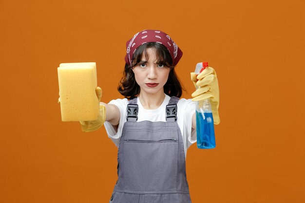 均一なゴム手袋とバンダナを身に着けている厳格な若い女性クリーナーは、オレンジ色の背景で隔離されたカメラに向かってスポンジとクレンザーを伸ばしているカメラを見て