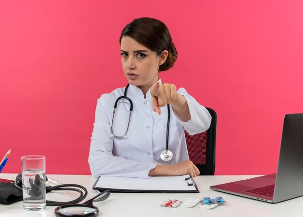 의료 가운과 청진기를 착용하고 의료 도구와 노트북이 분홍색 벽에 고립 된 책상에 손을 넣어 책상에 앉아 엄격한 젊은 여성 의사