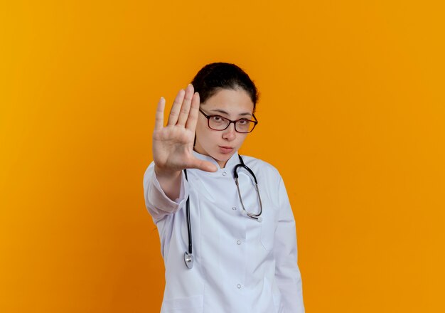 Strict young female doctor wearing medical robe and stethoscope with glasses showing stop gesture isolated