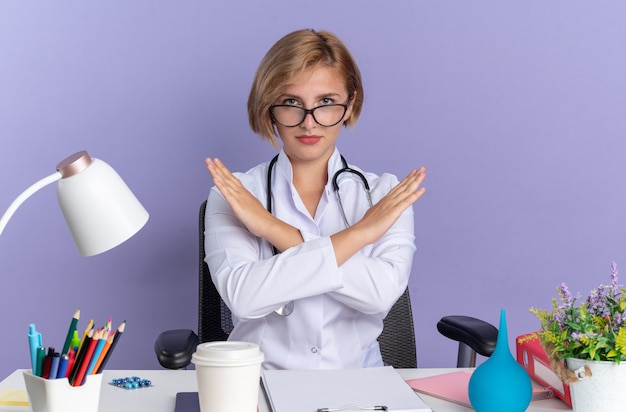 Rigorosa giovane dottoressa che indossa accappatoio medico con stetoscopio e occhiali si siede al tavolo con strumenti medici che mostrano gesto di non isolato su sfondo blu