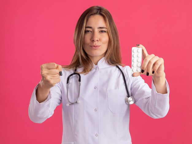 聴診器が丸薬を保持し、ピンクの壁に隔離された拳を差し出す医療ローブを身に着けている厳格な若い女性医師