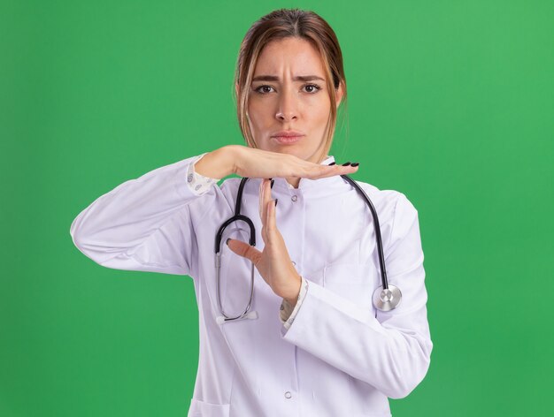 Strict young female doctor wearing medical robe with stethoscope showing timeout gesture isolated on green wall