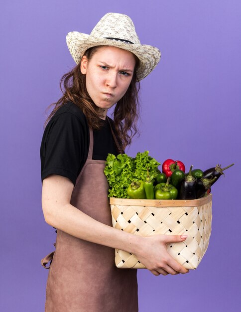 Strict young female gardener wearing gardening hat holding vegetable basket