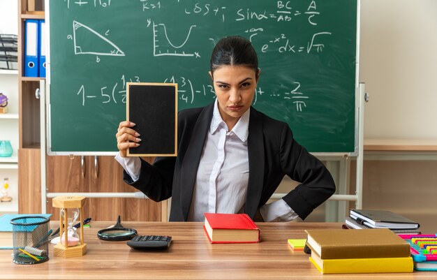 strict young female teacher sits at table with school supplies holding mini blackboard in classroom