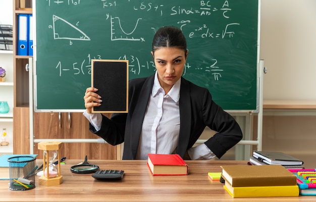 厳格な若い女性教師が教室でミニ黒板を保持している学用品とテーブルに座っています