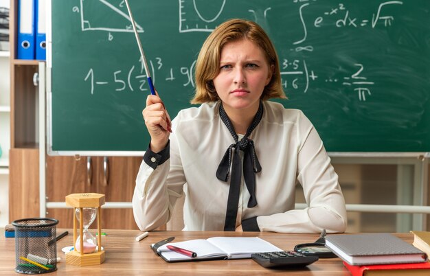 strict young female teacher sits at table with school supplies holding pointer stick in classroom