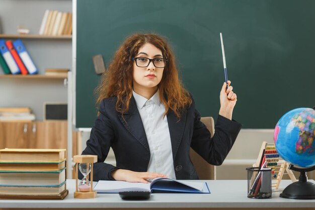 strict young female teacher wearing glasses points at blackboard with pointer sitting at desk with school tools in classroom