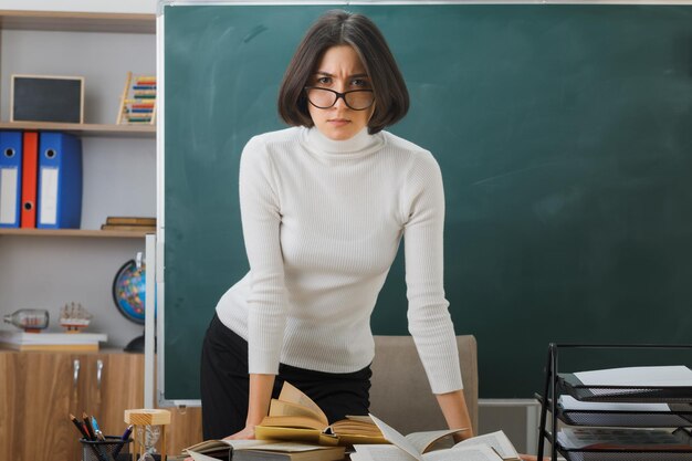 strict young female teacher wearing glasses standing in front of blackboard in classroom