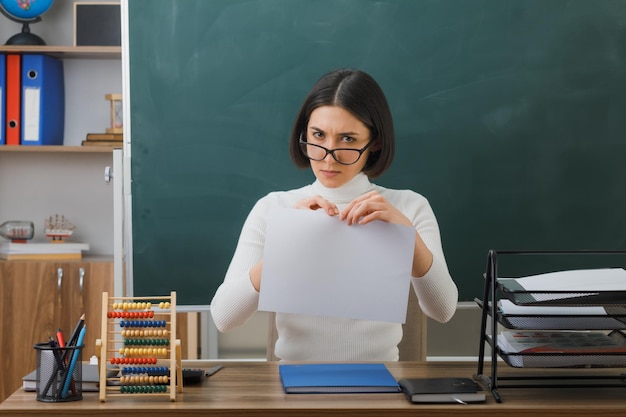 strict young female teacher wearing glasses tear paper sitting at desk with school tools on in classroom