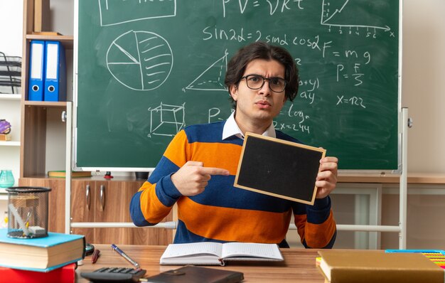 strict young geometry teacher wearing glasses sitting at desk with school supplies in classroom showing mini blackboard pointing at it looking at front