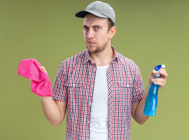 Strict young guy cleaner wearing cap holding cleaning agent with rag isolated on olive green wall