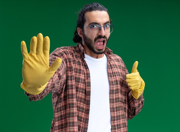 Строгий молодой красивый уборщик в футболке и перчатках, подняв большой палец вверх, показывает жест стоп, изолированный на зеленой стене