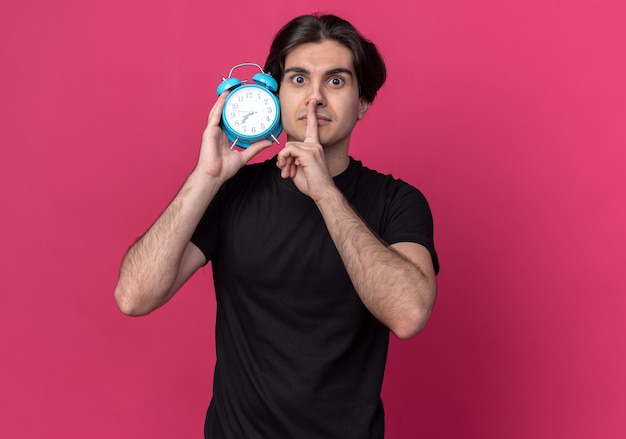 Strict young handsome guy wearing black t-shirt holding alarm clock around ear showing silence gesture isolated on pink wall