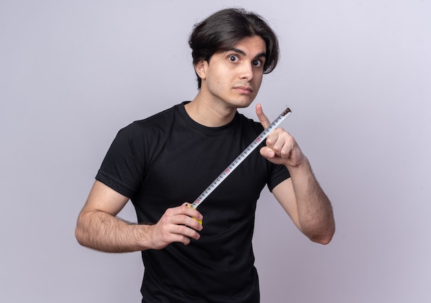 Strict young handsome guy wearing black t-shirt holding tape measure isolated on white wall