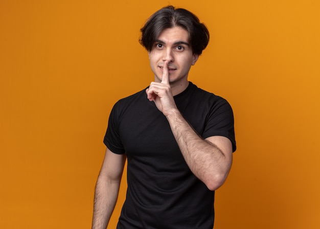 Strict young handsome guy wearing black t-shirt showing silence gesture isolated on orange wall