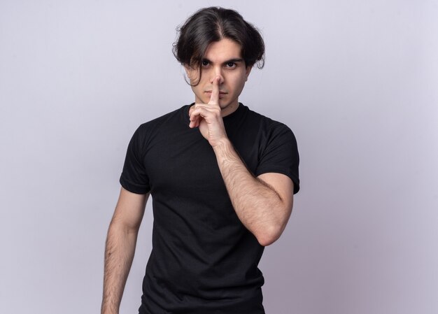 Strict young handsome guy wearing black t-shirt showing silence gesture isolated on white wall