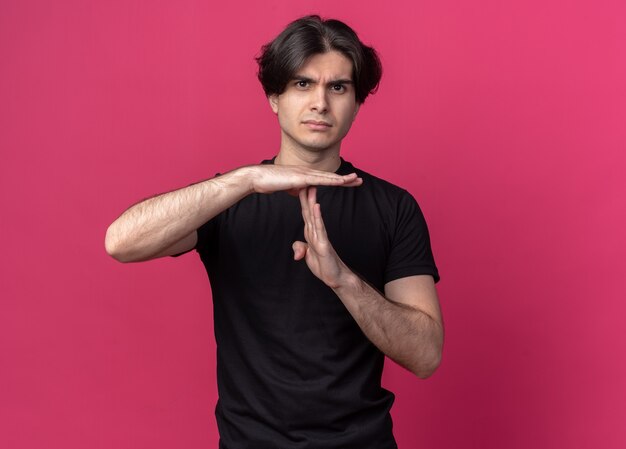 ピンクの壁に分離されたタイムアウトジェスチャーを示す黒いTシャツを着ている厳格な若いハンサムな男