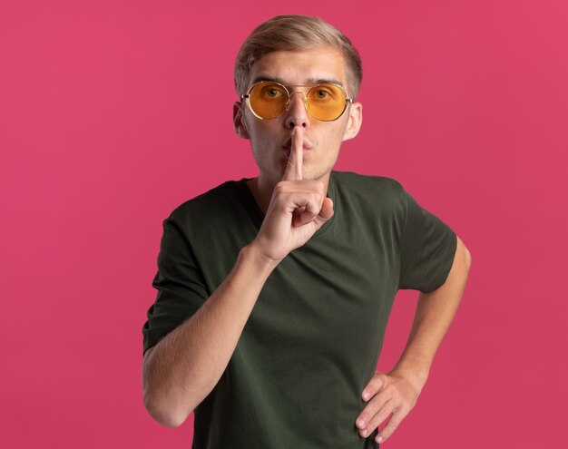 Free photo strict young handsome guy wearing green shirt and glasses showing silence gesture putting hand on hip isolated on pink wall