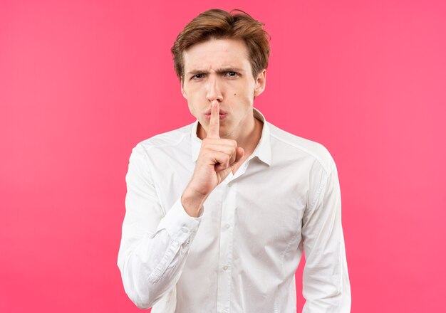 Strict young handsome guy wearing white shirt showing silence gesture isolated on pink wall