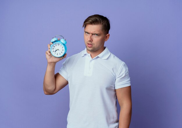 Strict young handsome male student holding alarm clock isolated on blue