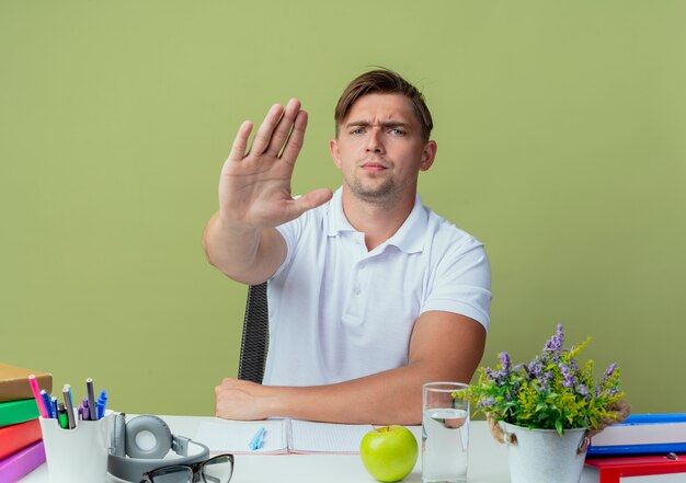Strict young handsome male student sitting at desk with school tools showing stop gesture on olive green