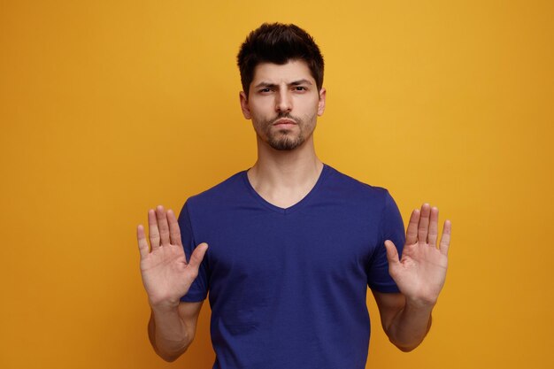 Strict young handsome man looking at camera doing stop gesture on yellow background