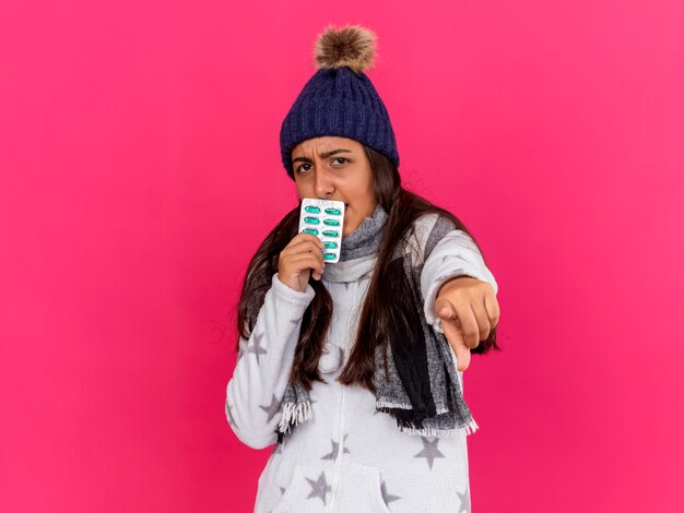 Strict young ill girl wearing winter hat with scarf holding pills around mouth showing you gesture isolated on pink