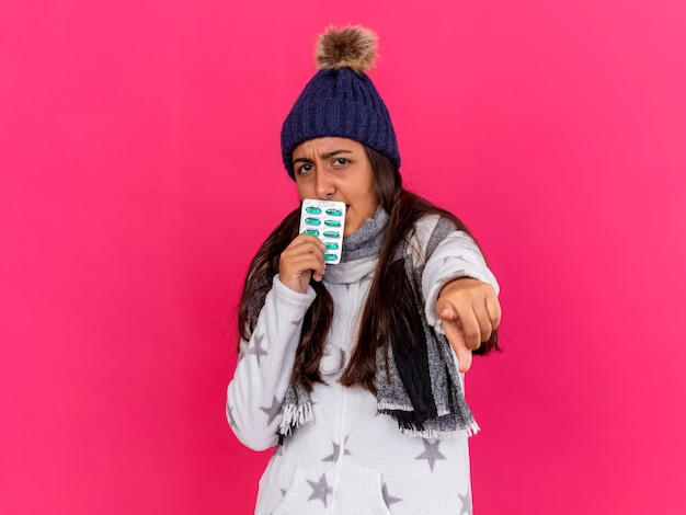 Строгая молодая больная девушка в зимней шапке с шарфом держит таблетки вокруг рта, показывая вам жест, изолированный на розовом