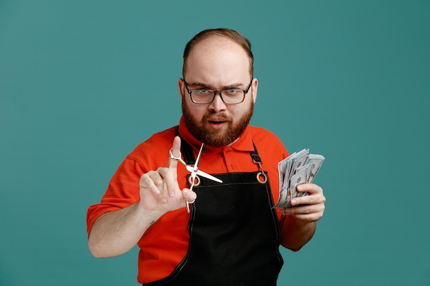 Free photo strict young male barber wearing glasses red shirt and barber apron holding money showing scissors looking at camera isolated on blue background