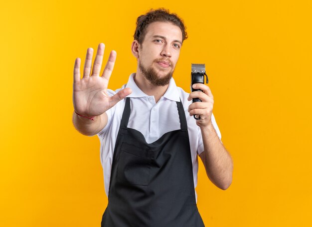 Строгий молодой мужчина-парикмахер в униформе, держащий машинку для стрижки волос, показывая жест остановки, изолированный на желтом фоне