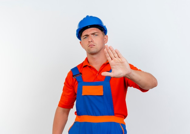 Бесплатное фото Строгий молодой мужчина-строитель в униформе и защитном шлеме, показывающий жест стоп на белом