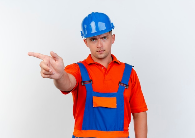 Strict young male builder wearing uniform and safety helmet points at side on white