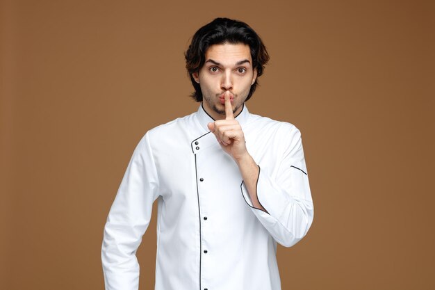 strict young male chef wearing uniform looking at camera showing silence gesture isolated on brown background