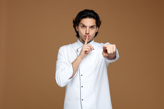 strict young male chef wearing uniform looking and pointing at camera showing silence gesture isolated on brown background