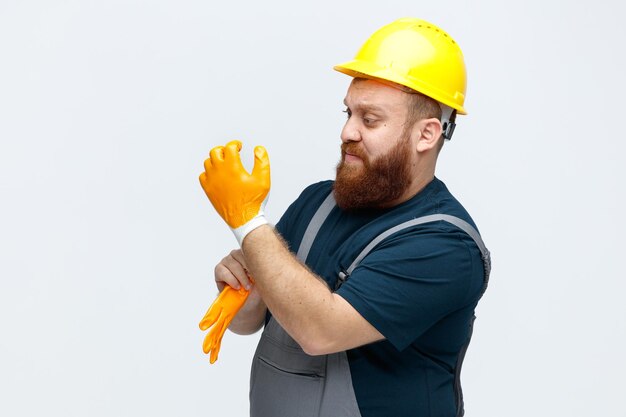 Strict young male construction worker wearing safety helmet and uniform standing in profile view wearing safety gloves looking at his own hand isolated on white background with copy space