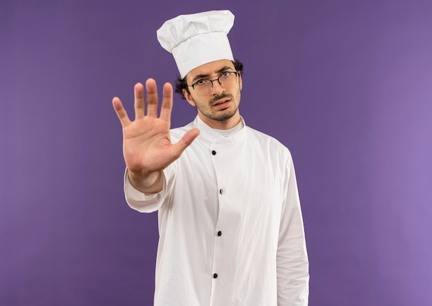 Strict young male cook wearing chef uniform and glasses showing stop gesture on purple