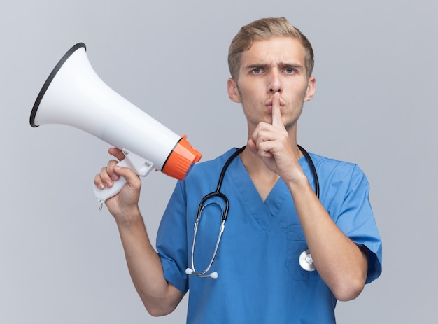 Free photo strict young male doctor wearing doctor uniform with stethoscope holding loudspeaker showing silence gesture isolated on white wall