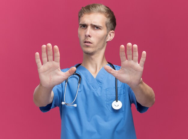 Строгий молодой мужчина-врач в униформе врача со стетоскопом, протягивая руки к камере, изолированной на розовой стене